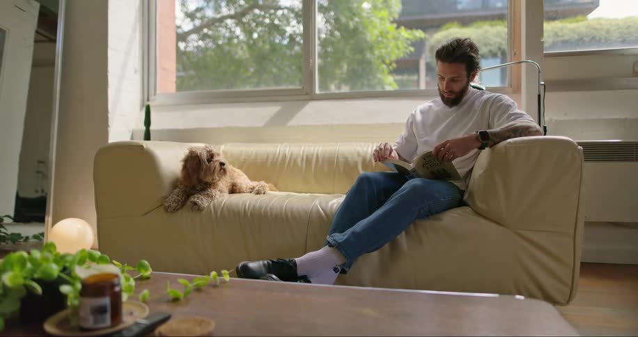 A man is at home with his dog while sitting on a sofa flipping through the pages of a book in the daytime
