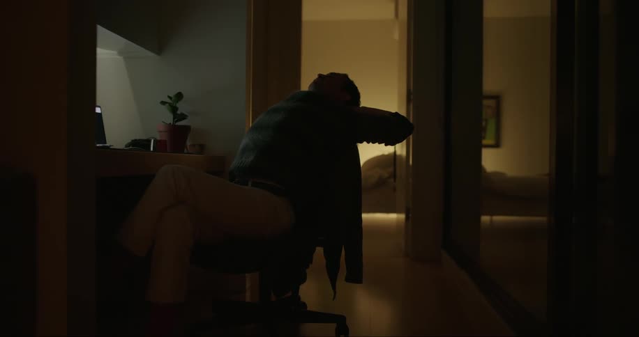 A man dressed in a cozy sweater and pants sits in an armchair diligently working on his laptop during the late hours in the dimly lit ambiance of his home at night