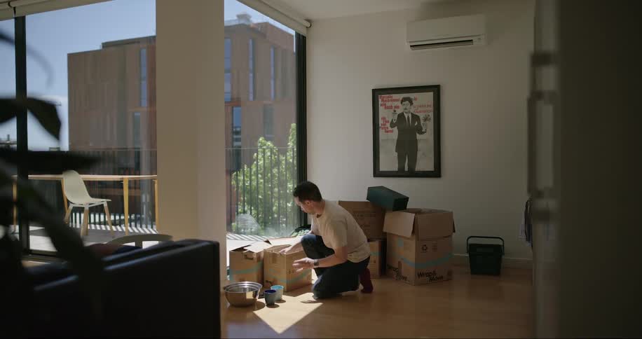 A man kneeling on the floor of a living room unpacks kitchenware from cardboard boxes