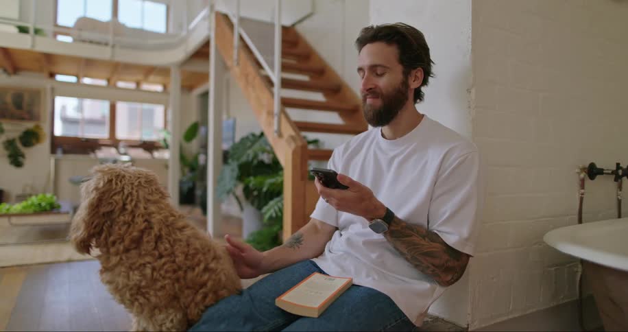A man putting on a white top and blue jean is petting his dog while operating his phone