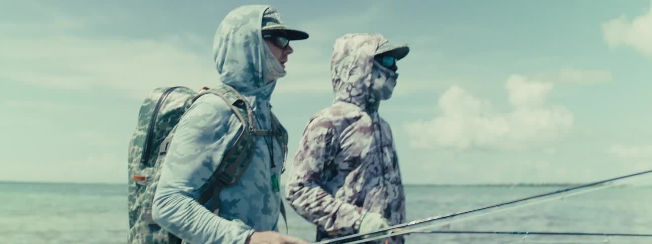 Two fishers dressed in blue and gray camouflage sporting gear standing talking pointing and looking out over a large body of water with fishing poles in their hands