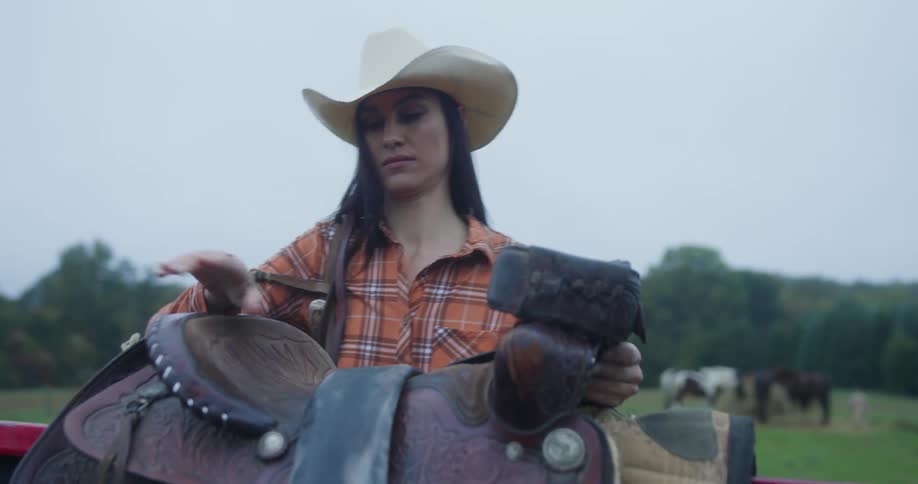 A woman wearing a cowboy hat places a horse saddle on a fence