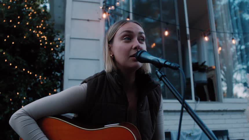 Woman seated in her backyard on a small stage playing the guitar and singing into a microphone