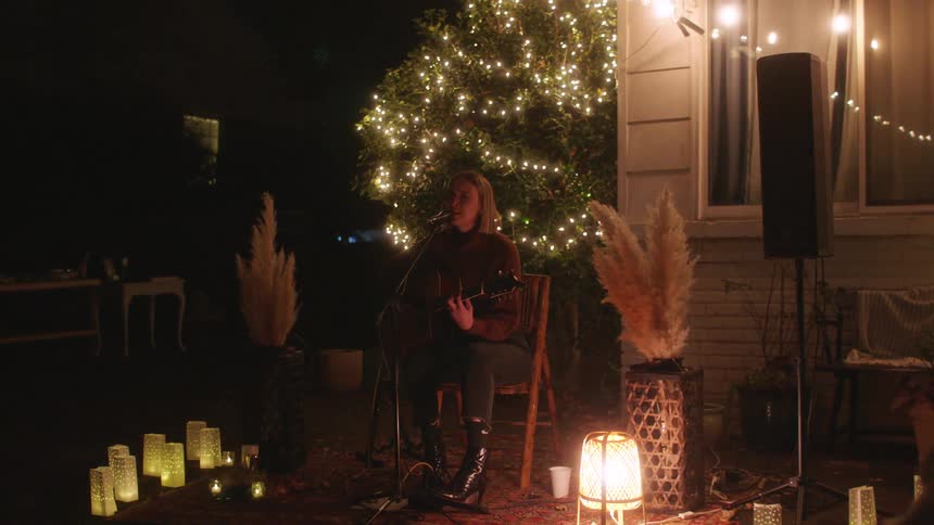 A seated woman with guitar strums and sings at an evening performance lit by paper lanterns