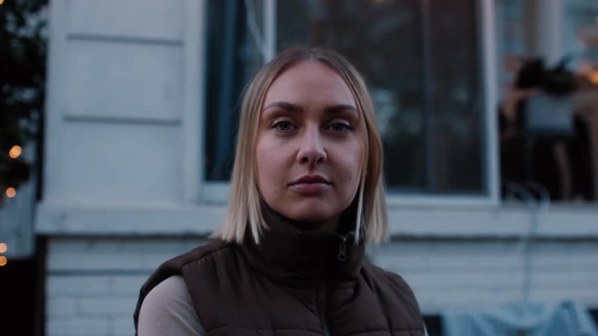 Blonde woman sitting outside the house in a chair in front of the window wearing a brown jacket