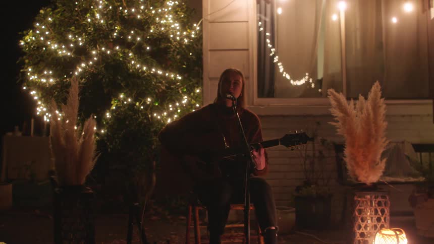 Woman sitting on decorated patio at night sings and plays the guitar