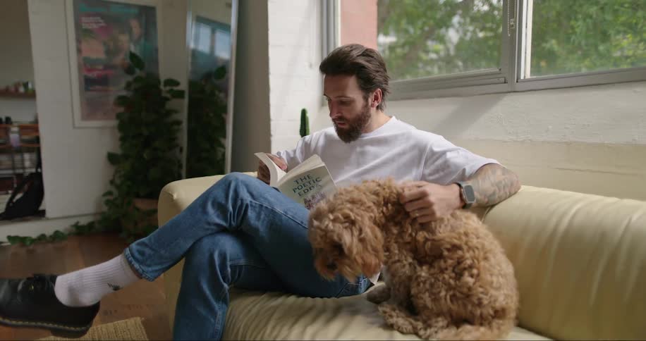 Man sitting on the sofa reading a book and petting his dog