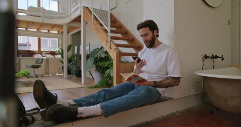 A man sitting is playing with a cell phone and smiles and then starts reading the book that is in his other hand