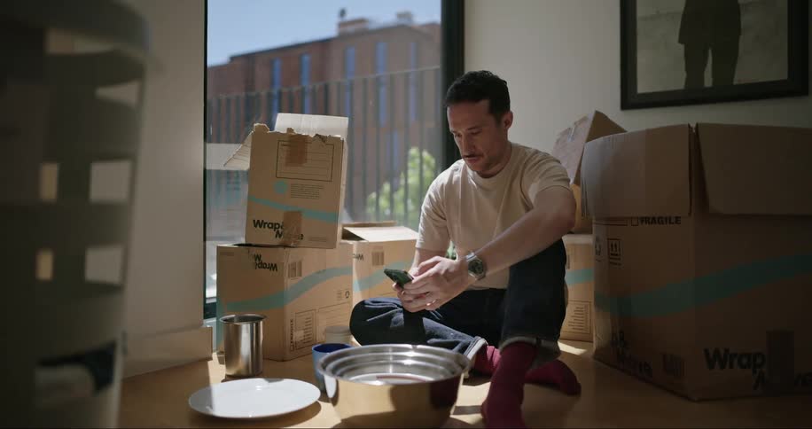 A man sits by moving boxes typing on his phone