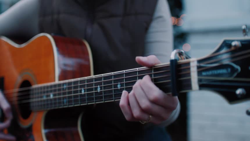 A woman sings intoa microphone and her fingers move to hold the strings on her guitar as she also plays in an outdoor setting at dusk