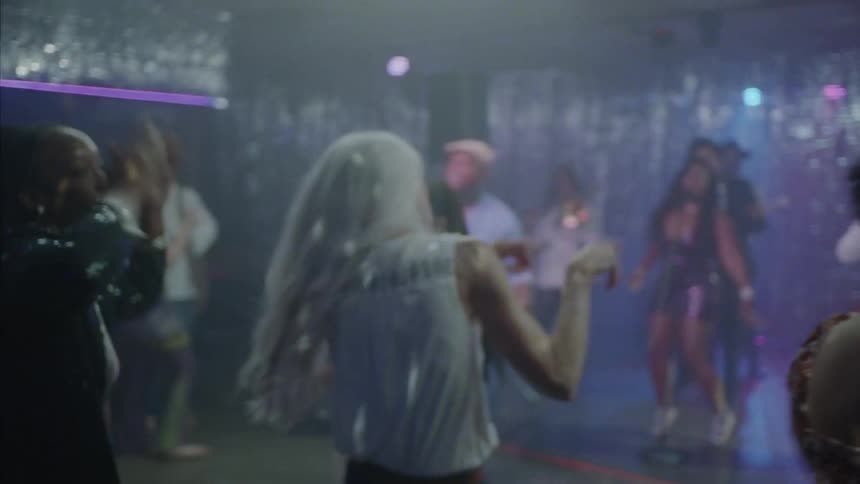 Grayhaired woman dancing in the middle of the dance floor with other people dancing around her at a dance club in front of a stage with musicians