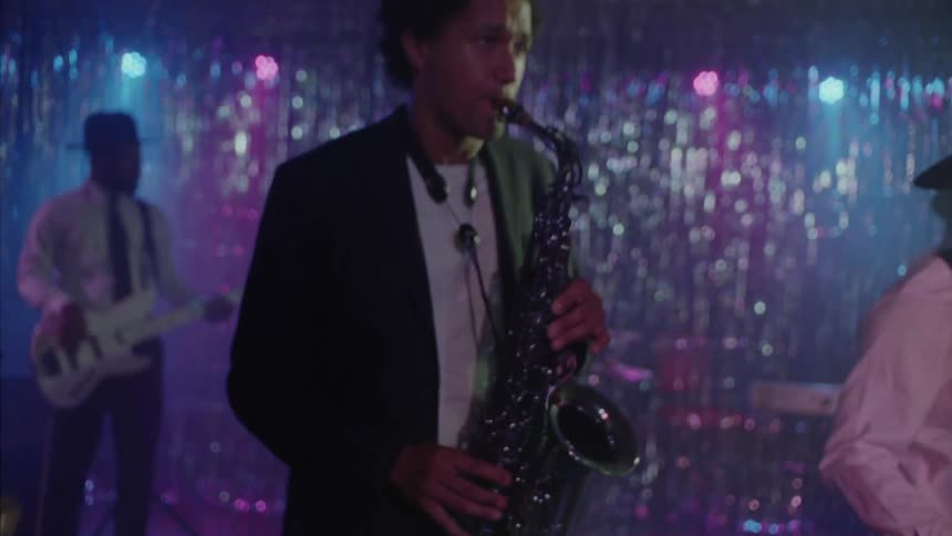 A woman with gray hair standing joyfully in front of a band at a nightclub with musicians playing various instruments on the stage including a saxophone