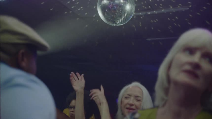 A woman with long blond hair dances around with others and they are all moving their arms around and clapping in a dark room under a disco light spinning with white lights on ceiling
