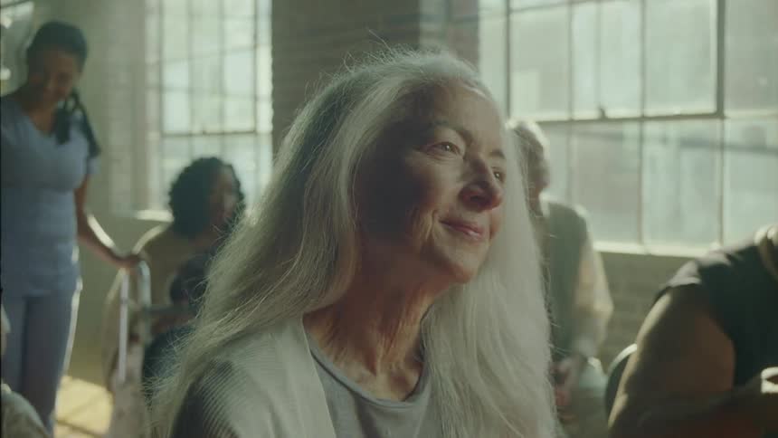 Portrait of an elderly woman sitting in a room with other people moving their heads and with a happy facial expression