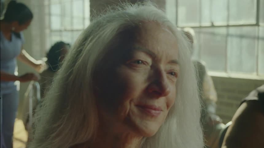 Old lady dances with her head in a room while seated in the midst of people during the day