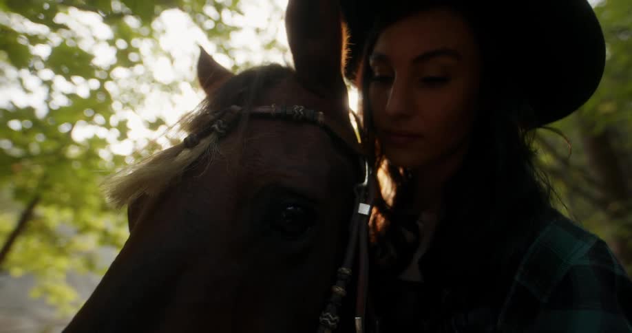 Woman in a cowboy hat gently stroking the face of a horse in a sunlit area surrounded by trees