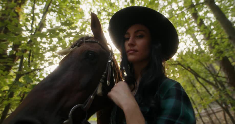 Woman wearing a cowboy hat standing next to a horse caressing its face in a forest