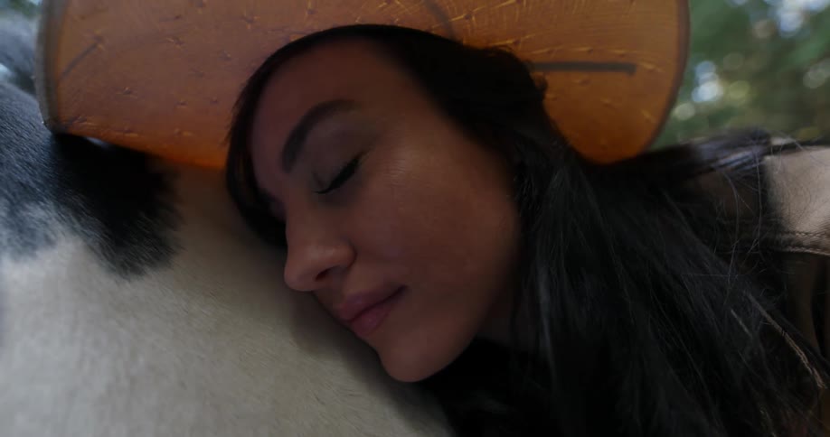 Woman with a sun hat a dark hair and a leather jacket cuddles a horse