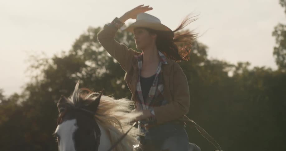 Young woman in jeans and a cowboy hat canters on a brown and white spotted horse