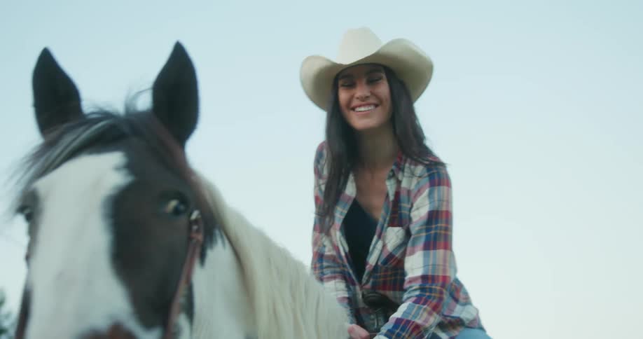 Woman wearing a cowboy hat a plaid shirt and jeans sits on horseback and pets it while laughing