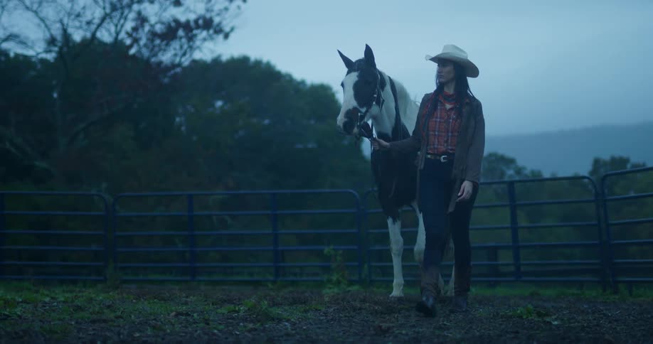 A woman is walking with her horse on a ranch in the mountains at dusk