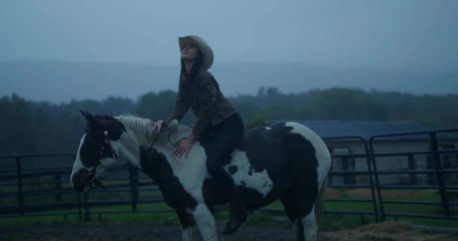 A woman dressed in cowgirl attire atop a horse on a farm under a cloudy sky