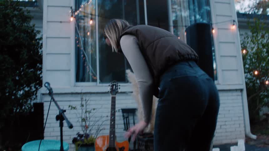 A young female adult smiling and talking bends down and moves guitar chords around on a rug with a stool set up behind a house with lights strung up for a small backyard performance in the fall at dusk