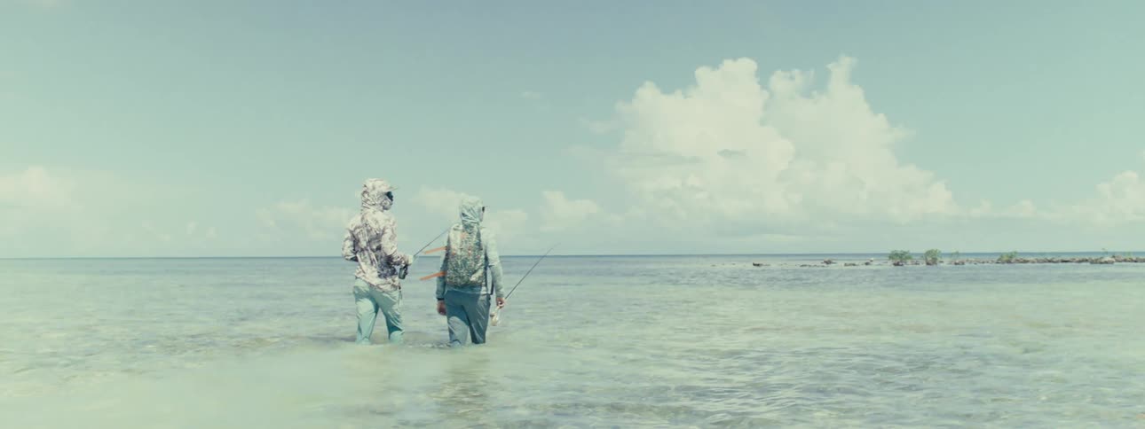 Two people walk in the ocean with the water at their knees carrying fishing rods