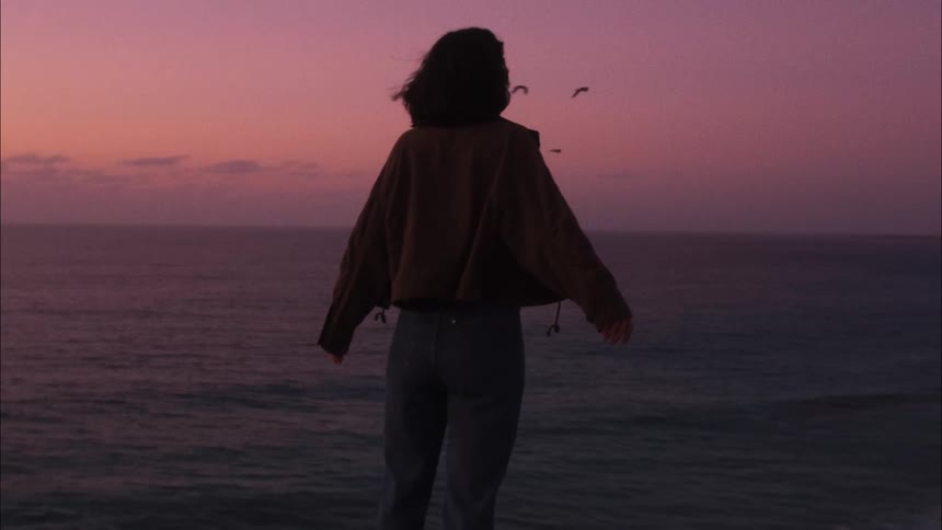 A woman in a brown jacket standing on the edge of a cliff taking in the sunset view of the sea ahead