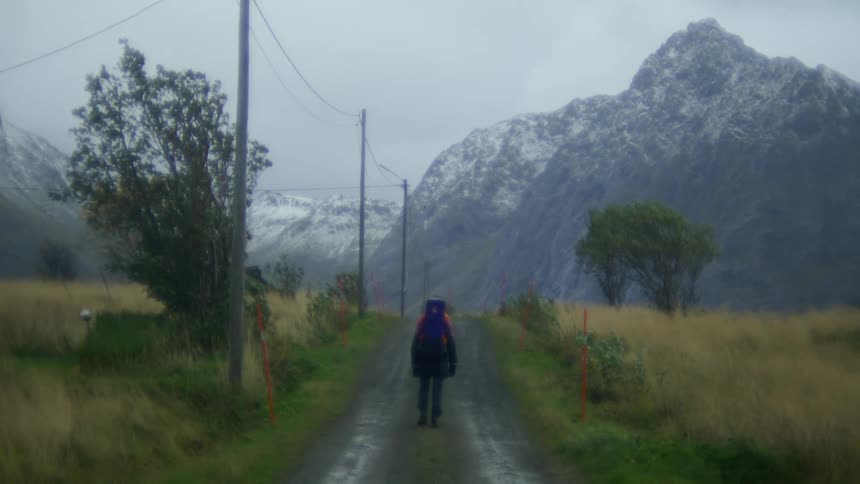 Hiker carrying child walks along dirt road between pastures and toward a snowy mountain