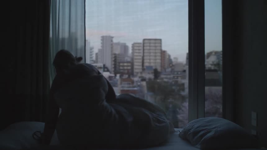 A woman wrapped in a blanket in bed watching the city through the window