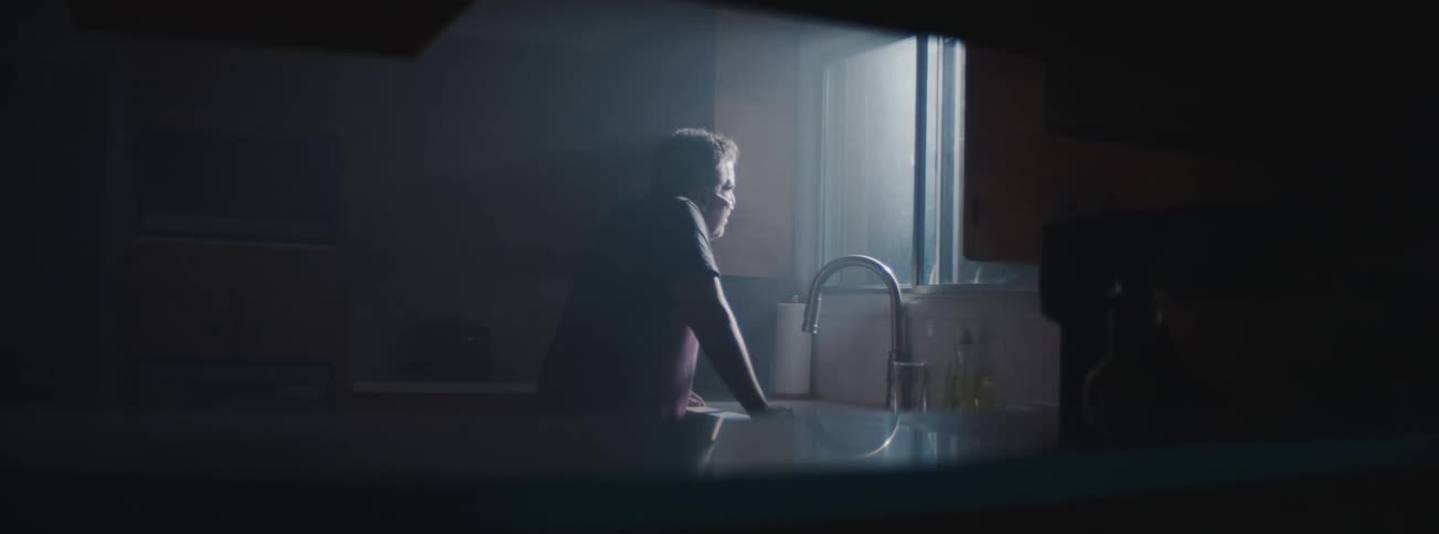 Senior old man walking slowly through the kitchen leaning on the sink and looking out the window