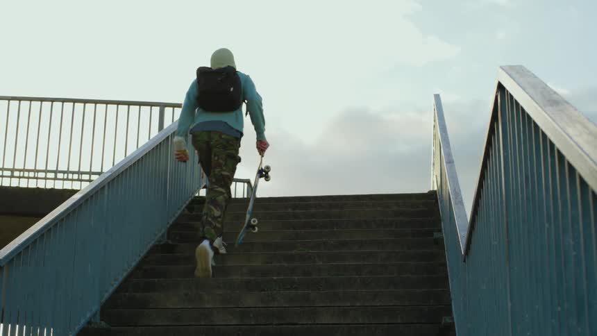 Young skateboarder with a backpack beanie and holding a skateboard climbing the stairs of a footbridge on a cloudy day
