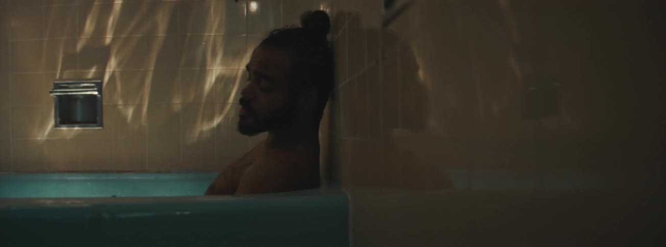A sad man with a beard is lying in a bathtub taking a shower