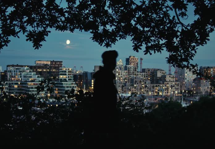 A man walking in the countryside is silhouetted against the city buildings in the background during dusk