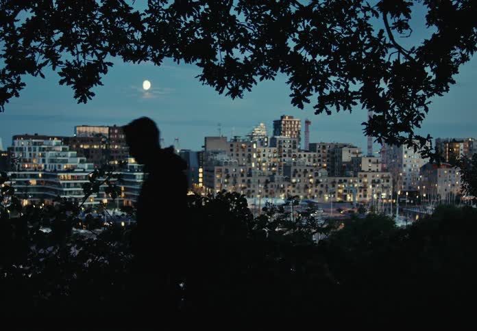 A man walking in the countryside is silhouetted against the city buildings in the background during dusk