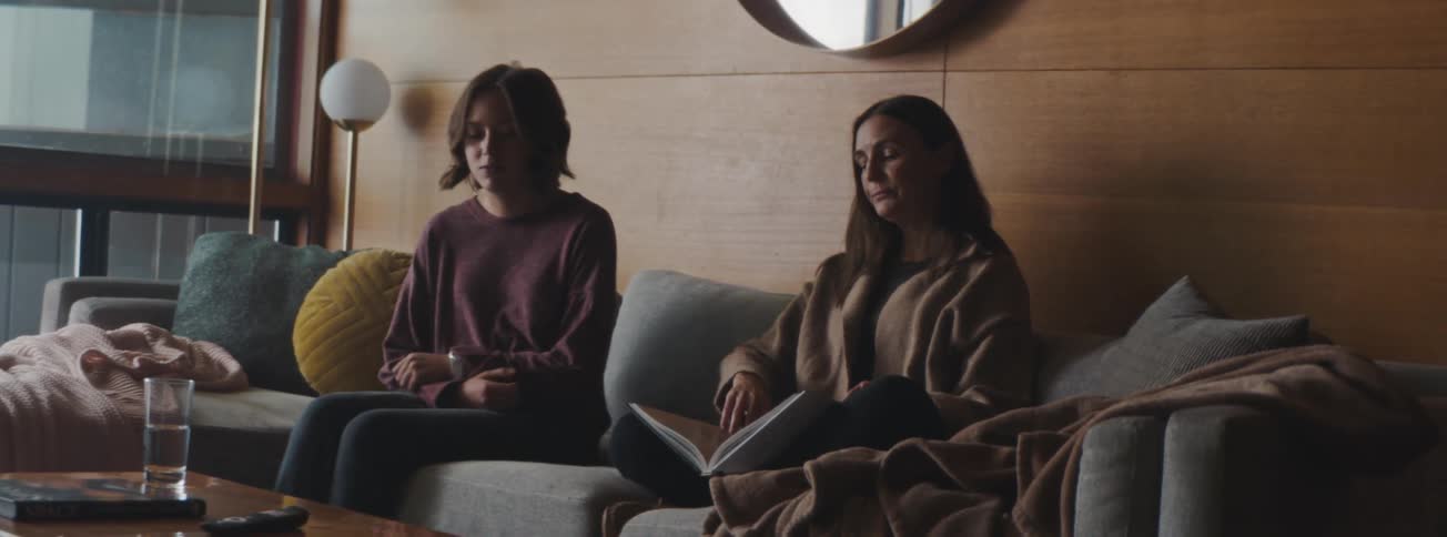 A woman sitting on the couch reading a book and her daughter comes join her and they start a conversation