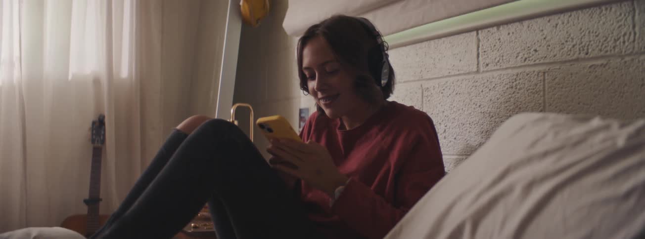 Female teen seated on the bed while listening to music and texting on mobile phone