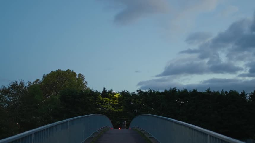 A person on a bicycle passing by another person walking on a narrow footbridge in the late afternoon