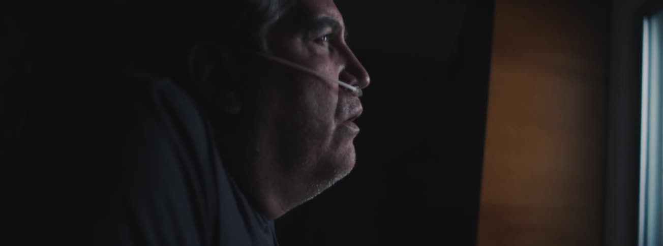 A senior man wearing a breathing aid approaches a bathroom sink and sighs while looking at the mirror