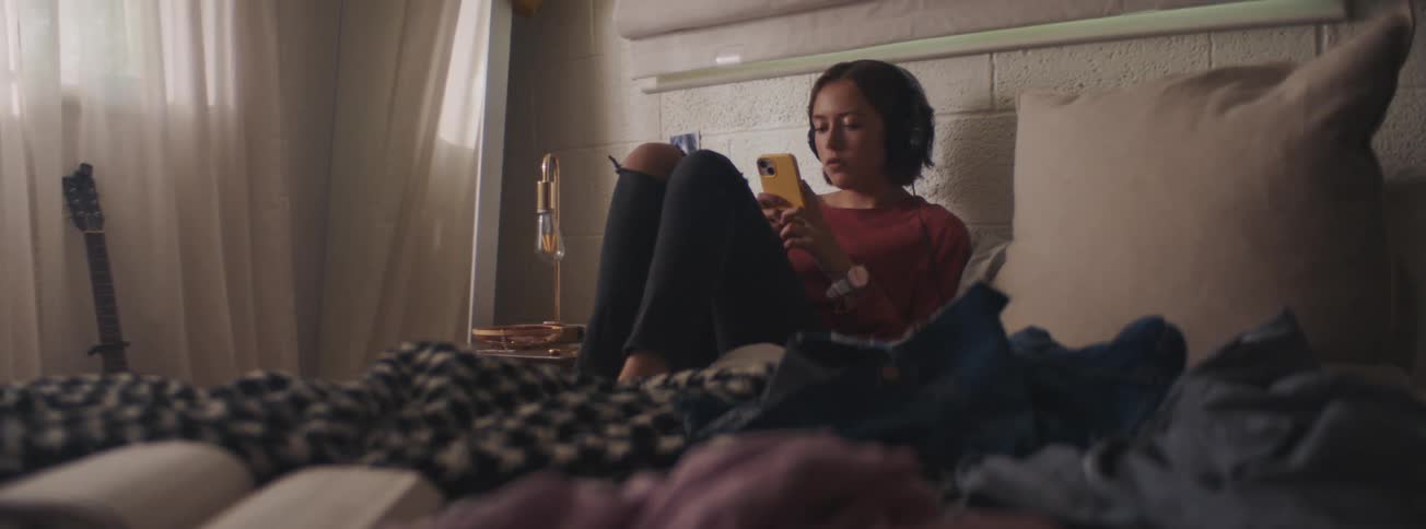 A woman is at home sitting in bed wearing headphones while using a smartphone during the day