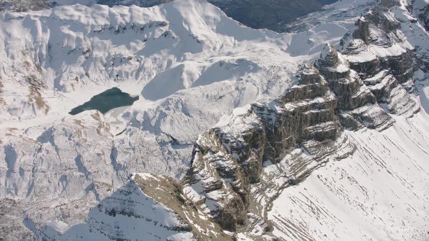 An aerial view of snow covered rocky mountains with a lake between two ridges on a sunny day