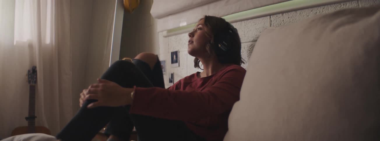A woman wearing ripped jeans and a red shirt sits on a bed immersed in music through her headphones