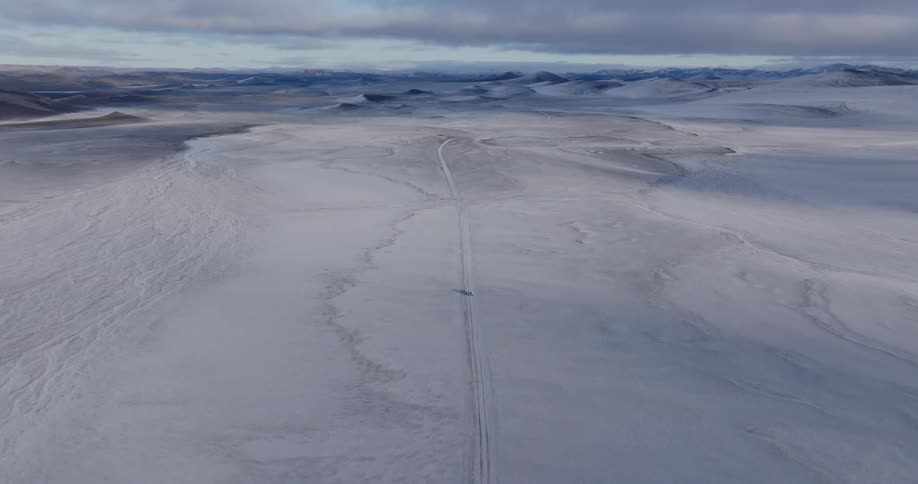 Aerial of a car driving down a flat ice road through a snow covered flat terrain towards artic mountain hills as the sun is just beginning to rise