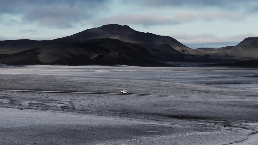 Vehicle drives along road through plains covered by dingy snow with hills in the background
