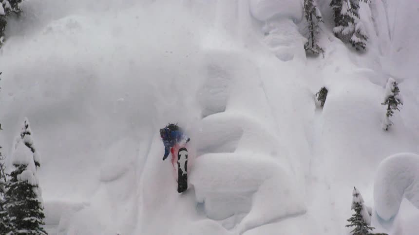 A snowboarder rides a pillow line in the powder in steep trees