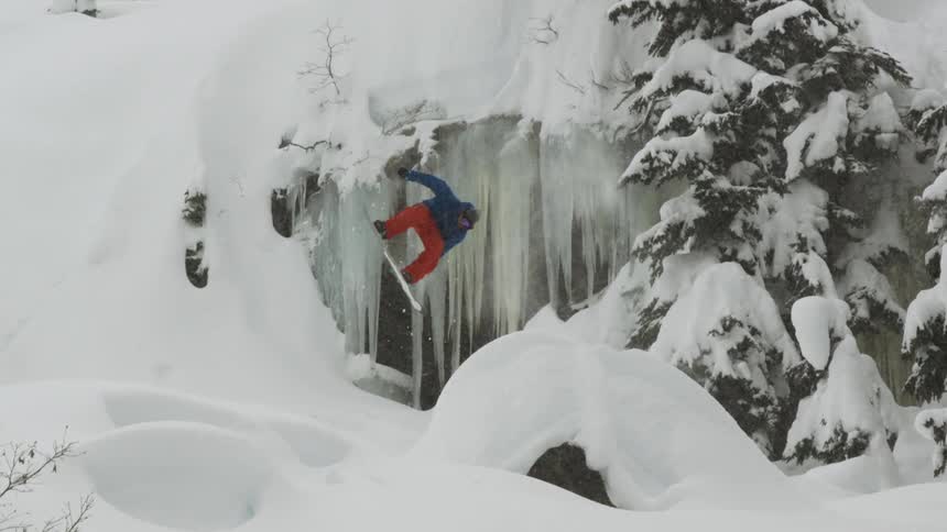 A snowboarder jumps and spins near a frozen waterfall