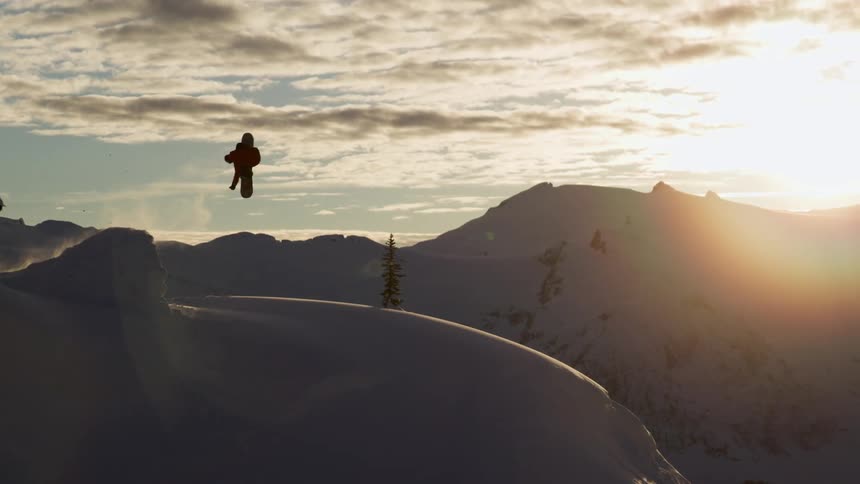 A snowboarder jumps and spins onto a steep slope as sun sets
