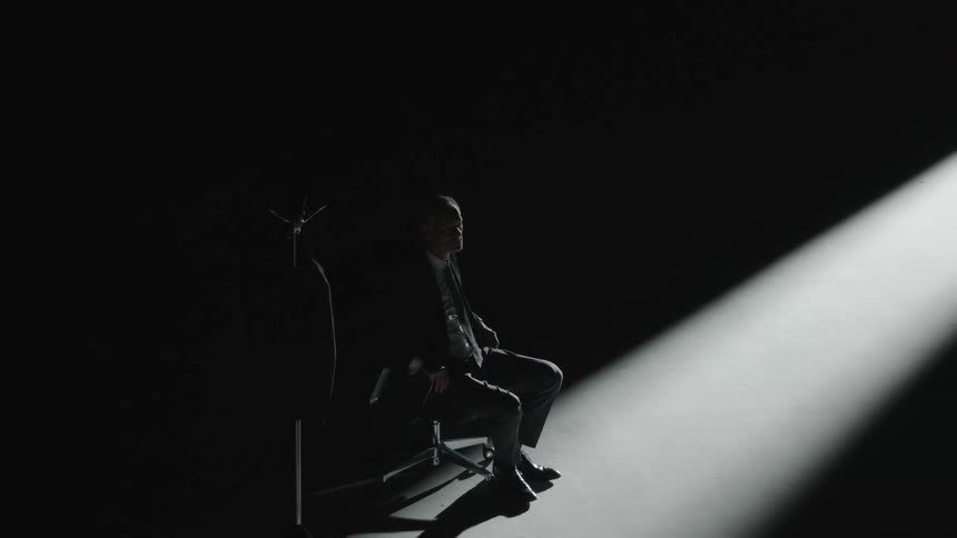 White man indoors on formal wear sitting down on an office chair in a dark room