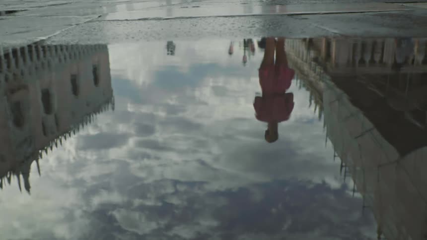 Puddle on a walkway with the reflection of a woman in a pink dress